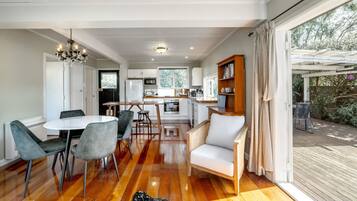 Una cocina moderna con armarios blancos, una zona de comedor con una mesa redonda y sillas, y una terraza de madera visible a través de un ventanal grande.