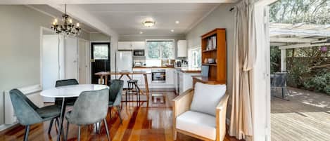 Une cuisine moderne avec des meubles blancs, un coin repas doté d’une table ronde et de chaises, et une terrasse en bois visible à travers une grande fenêtre.