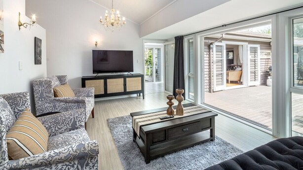 A modern living room with a large TV, patterned sofas, a dark coffee table, and a view of a patio with outdoor furniture. - Charming Character Retreat | 2BR, Private Deck & Year-Round Comfort (Martinborough)