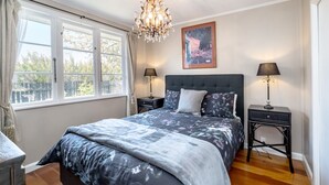 A bedroom with a large bed, two bedside tables, a chandelier, and a window with curtains. - Charming Character Retreat | 2BR, Private Deck & Year-Round Comfort (Martinborough)