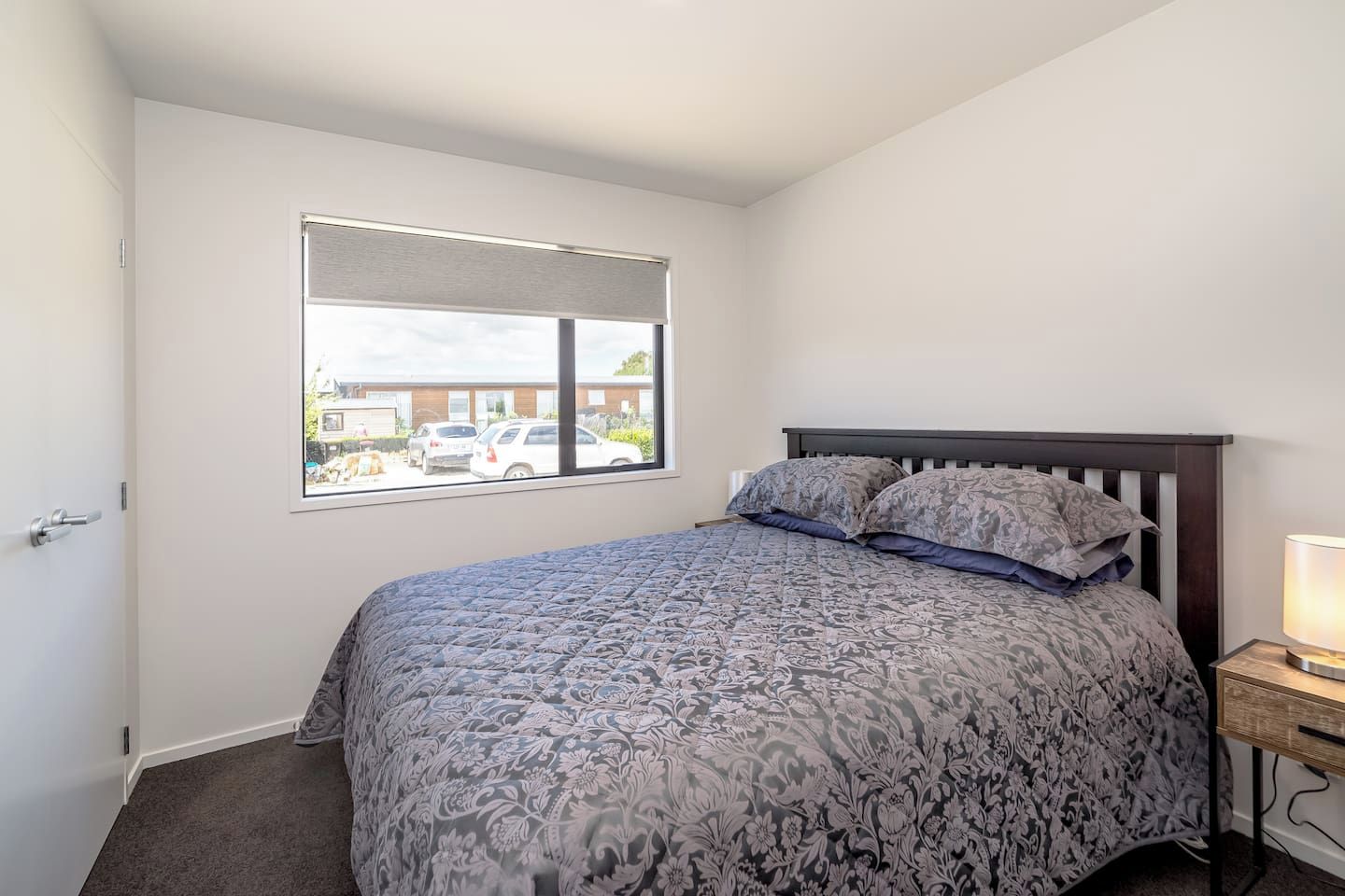 A bedroom with a bed, a nightstand, a lamp, a window, and a view of the outside.