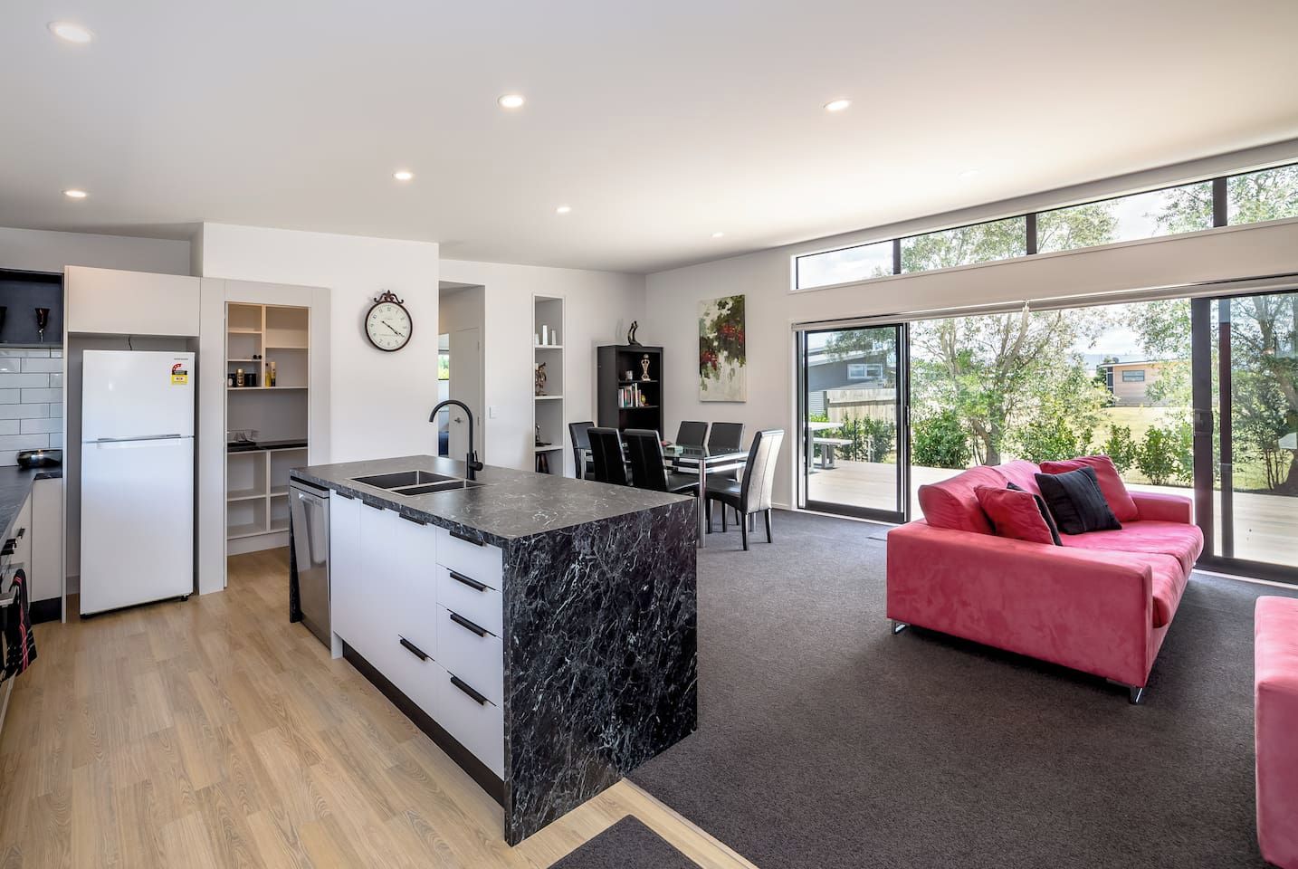 A modern kitchen with a marble island, a dining area with a red sofa, and a view through large windows.