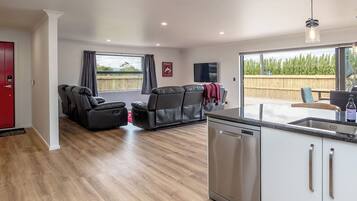 A modern living room with a sofa and a flat-screen TV, a kitchen with a dishwasher, and a view of a wooden deck.
