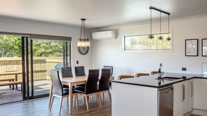 A modern kitchen with a central island, a dining area with chairs, and a large sliding glass door leading to an outdoor deck. - Broadway On New York (Martinborough)