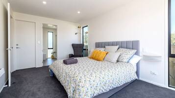 A modern bedroom with a large bed, a grey upholstered chair, a bedside table, and a view of the outdoors.