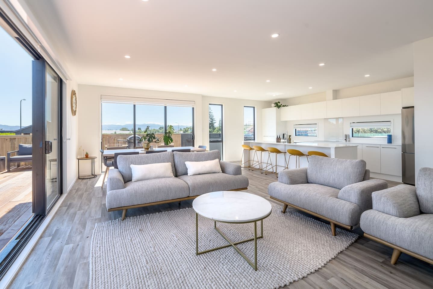 A modern living room with a sofa and coffee table, a kitchen with white cabinets, and large windows offering a view.