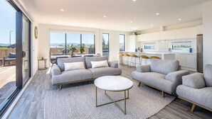 Ein modernes Wohnzimmer mit einer grauen Sofagarnitur, einem runden Couchtisch und einem großen Fenster mit Blick ins Freie.