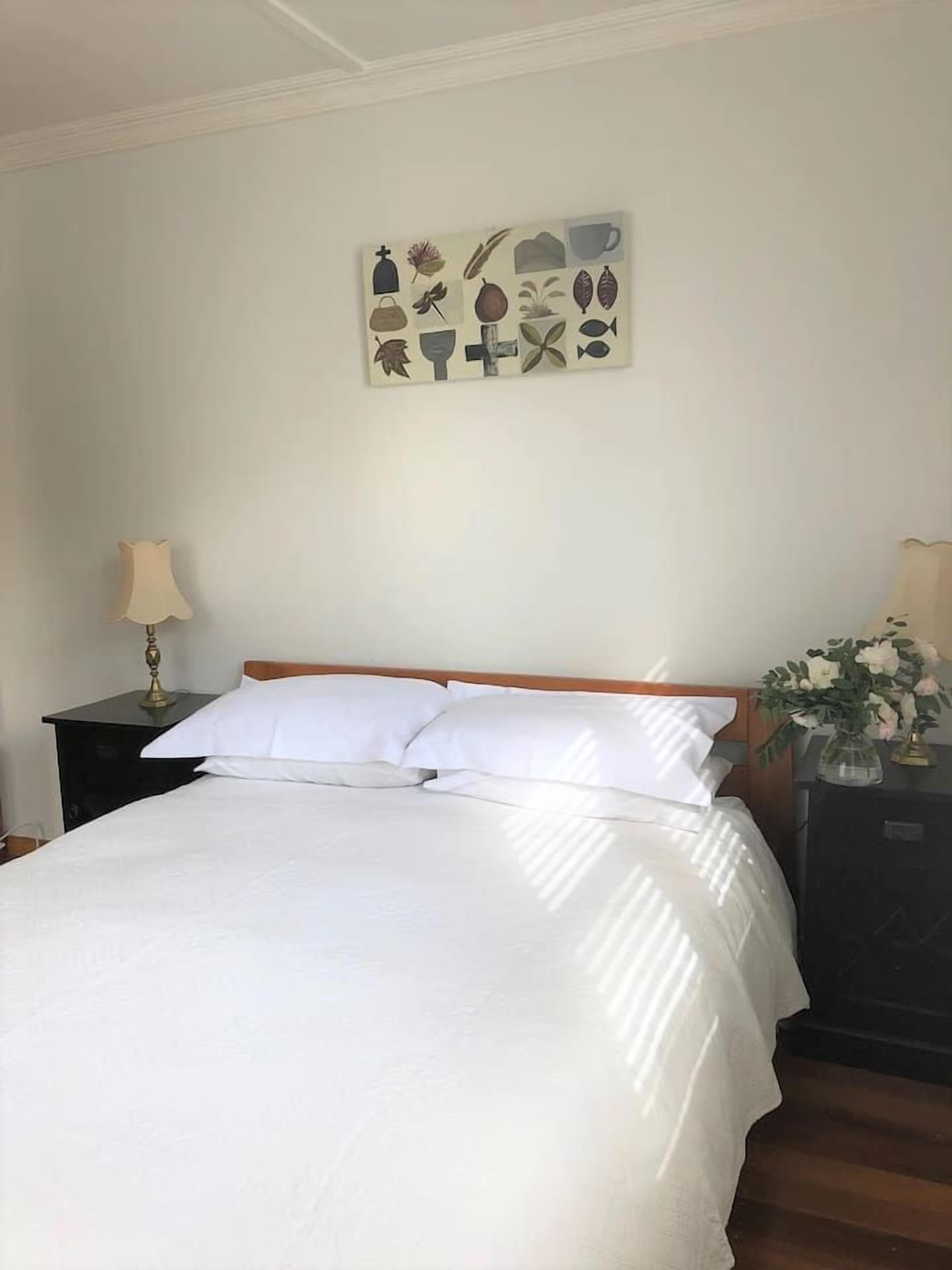 Un lit bien fait, avec du linge de lit blanc, une tête de lit en bois, une table de chevet avec une lampe, et une œuvre d’art accrochée au mur au-dessus du lit.