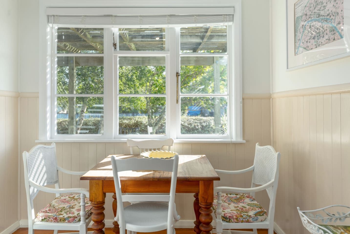 A dining area with a wooden table and four chairs, a window with a view of greenery, and a framed map on the wall.