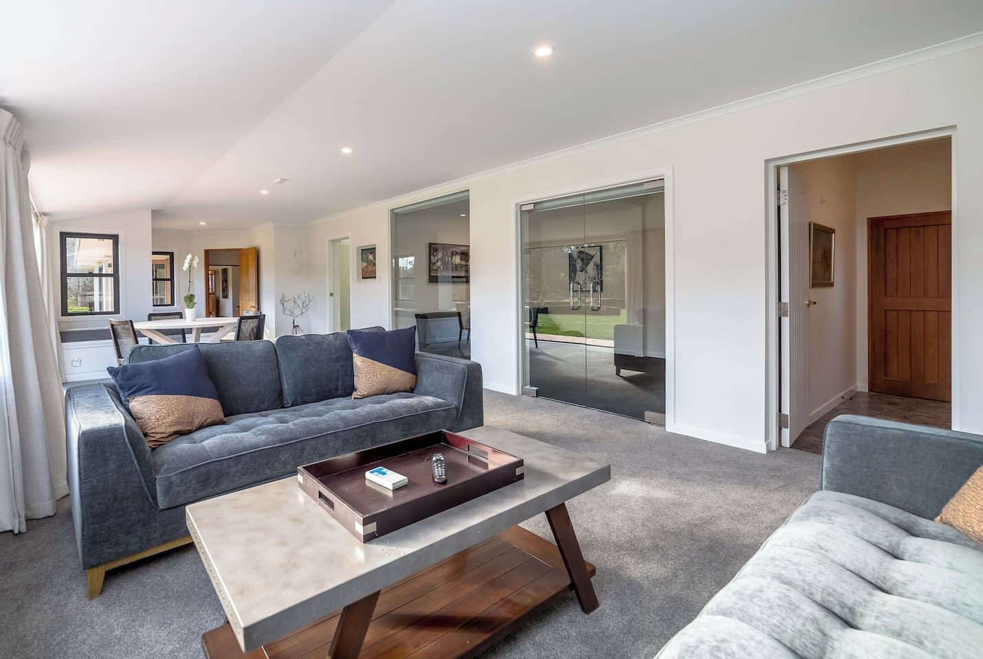 A modern living room with a grey sofa, a coffee table, and a view of the backyard through glass doors.