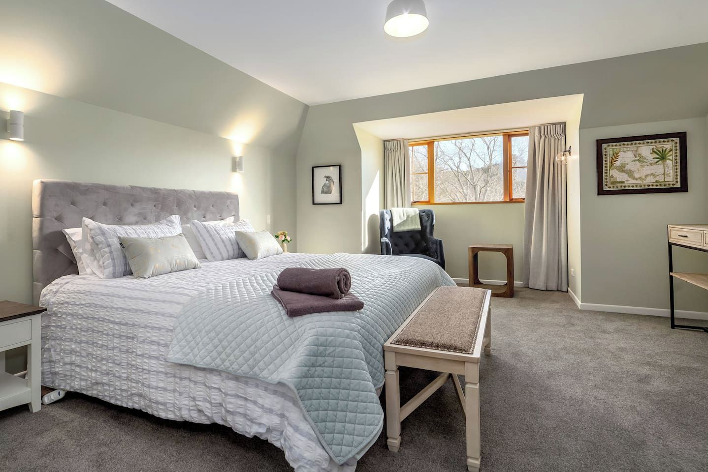 Une chambre à coucher avec un grand lit, des tables de chevet, une chaise, une petite table et une fenêtre avec des rideaux.