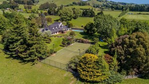 Vue aérienne d’une propriété comprenant un court de tennis, une grande maison et un espace vert environnant.