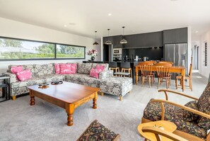 A modern living room with a sofa, coffee table, and chairs, a dining area with a table and chairs, a kitchen with appliances, and large windows offering a view. - Relax on the Deck, Martinborough (Martinborough)