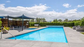 A rectangular swimming pool with a shaded area and lounge chairs, surrounded by a hedge and a wooden fence.