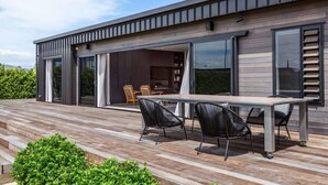 Uma casa moderna com um amplo deque, uma área de refeições com cadeiras e uma porta de vidro deslizante que dá para o interior.