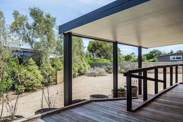 A covered outdoor deck with a wooden floor and black railings, surrounded by a garden with various plants and a gravel area.