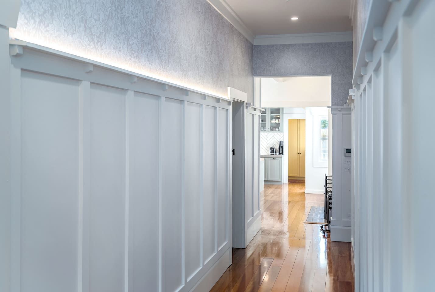 A hallway with white paneling, a kitchen area with cabinets, and a wooden floor.
