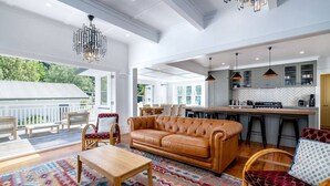 A spacious living room with a large sofa, a wooden coffee table, and a colorful rug. There is a kitchen area with bar stools and pendant lighting. - The Forest House, Martinborough (Pirinoa)