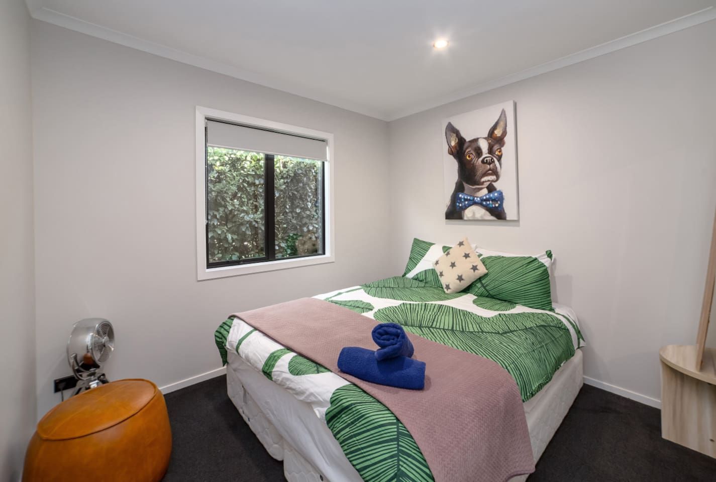 A bedroom with a bed, a window, a fan, a wooden stool, and a wall-mounted picture.