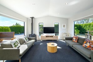 A spacious living room with a fireplace, a flat-screen TV, and a large window offering a view of the outdoors. - La Maison Noir, Martinborough (Martinborough)