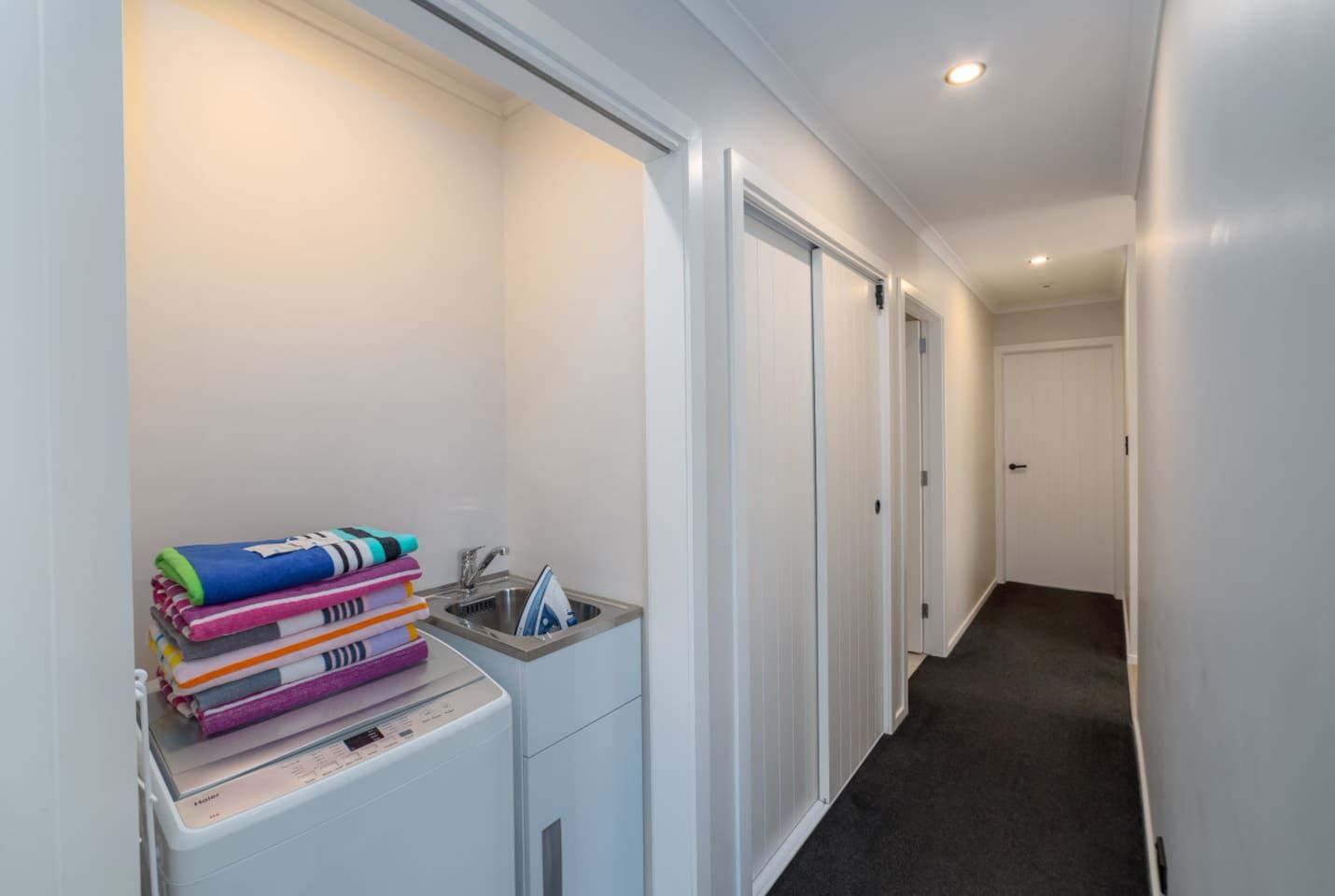 A narrow hallway with a laundry area on the left, featuring a stacked washer and dryer, a sink, and a pile of folded towels.