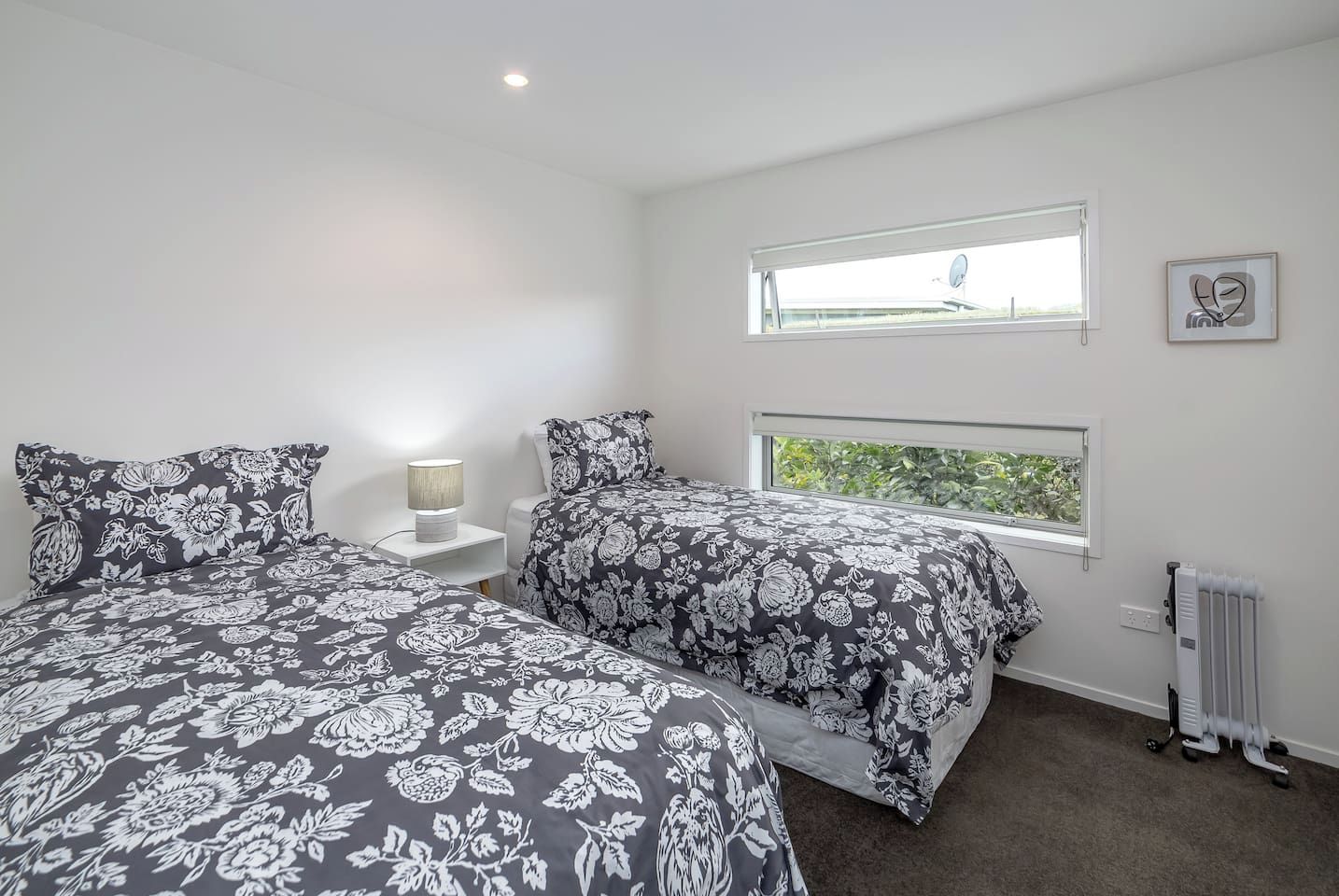 A bedroom with two beds, a nightstand, a lamp, a radiator, and a window with a view.