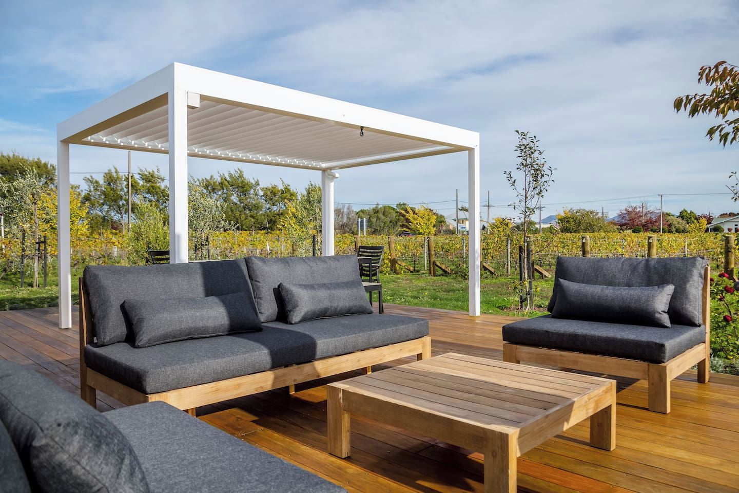 An outdoor seating area with a wooden deck, a white pergola, and a backdrop of a vineyard.