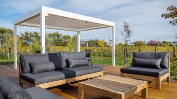 An outdoor seating area with a wooden deck, a white pergola, and a backdrop of a vineyard.