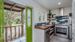 A compact kitchen with a gas stove, oven, and a sink. There is a window with a view of trees outside. - The Glade, Martinborough (Ruakōkoputuna)
