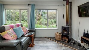 A cozy living room with a fireplace, a sofa with colorful cushions, and large windows overlooking greenery. - The Glade, Martinborough (Ruakōkoputuna)