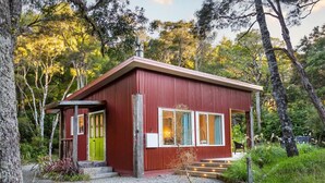 A small, red wooden cabin with a green door, a porch, and a chimney. - The Glade, Martinborough (Ruakōkoputuna)