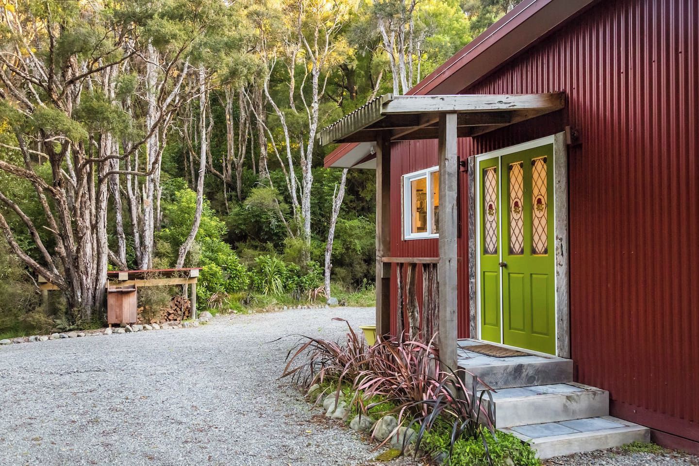 A rustic building with a green door, a wooden porch, and a gravel driveway leading to a wooden bench.