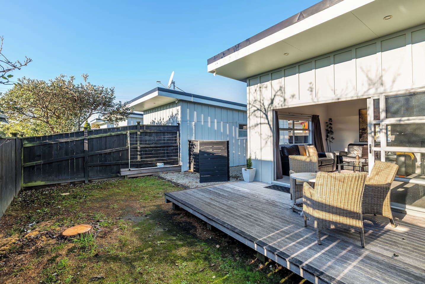A modern outdoor patio with a wooden deck, wicker furniture, and a glass door leading to an interior space.