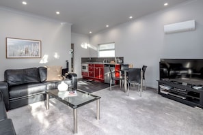 A modern living room with a leather sofa, a glass coffee table, a flat-screen TV, and a kitchen area with red cabinets and a dining table. - Two Four Cologne #3, Martinborough (Martinborough)