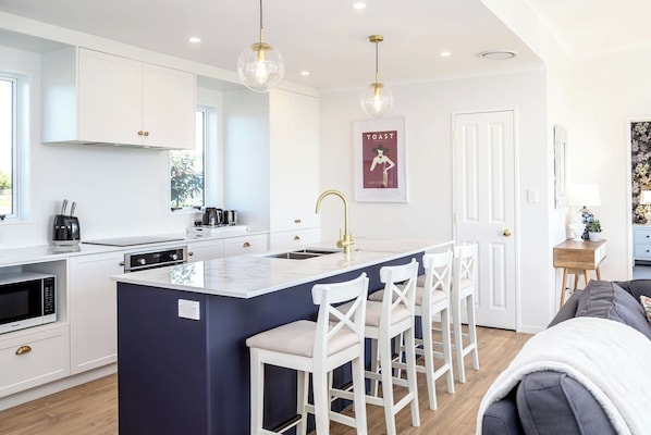 A modern kitchen with white cabinetry, a central island with bar stools, a built-in oven, and a pendant light. - Three on Wilson, Martinborough (Martinborough)