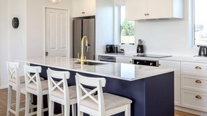 A modern kitchen with a central island, white cabinetry, and a large window providing natural light. - Three on Wilson | Modern 4BR, Seasonal Communal Pool Access & Walk to Town (Martinborough)