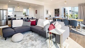 A modern living room with a grey sectional sofa, a dining area with a wooden table and chairs, and a kitchen in the background. - Three on Wilson | Modern 4BR, Seasonal Communal Pool Access & Walk to Town (Martinborough)