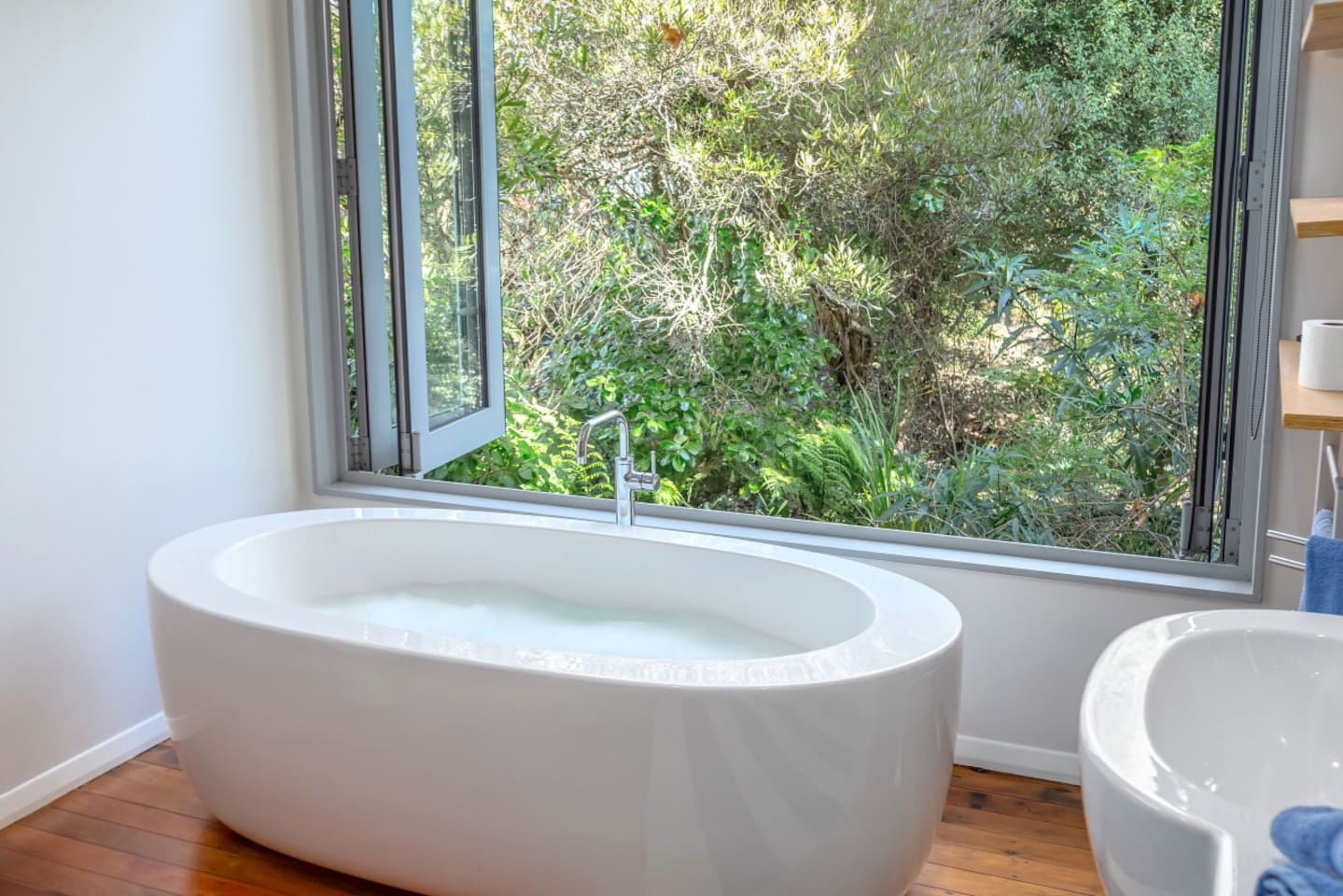 A modern bathroom with a freestanding bathtub, a window with a view of greenery, and a wooden floor.