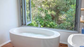 A modern bathroom with a freestanding bathtub, a window with a view of greenery, and a wooden floor.