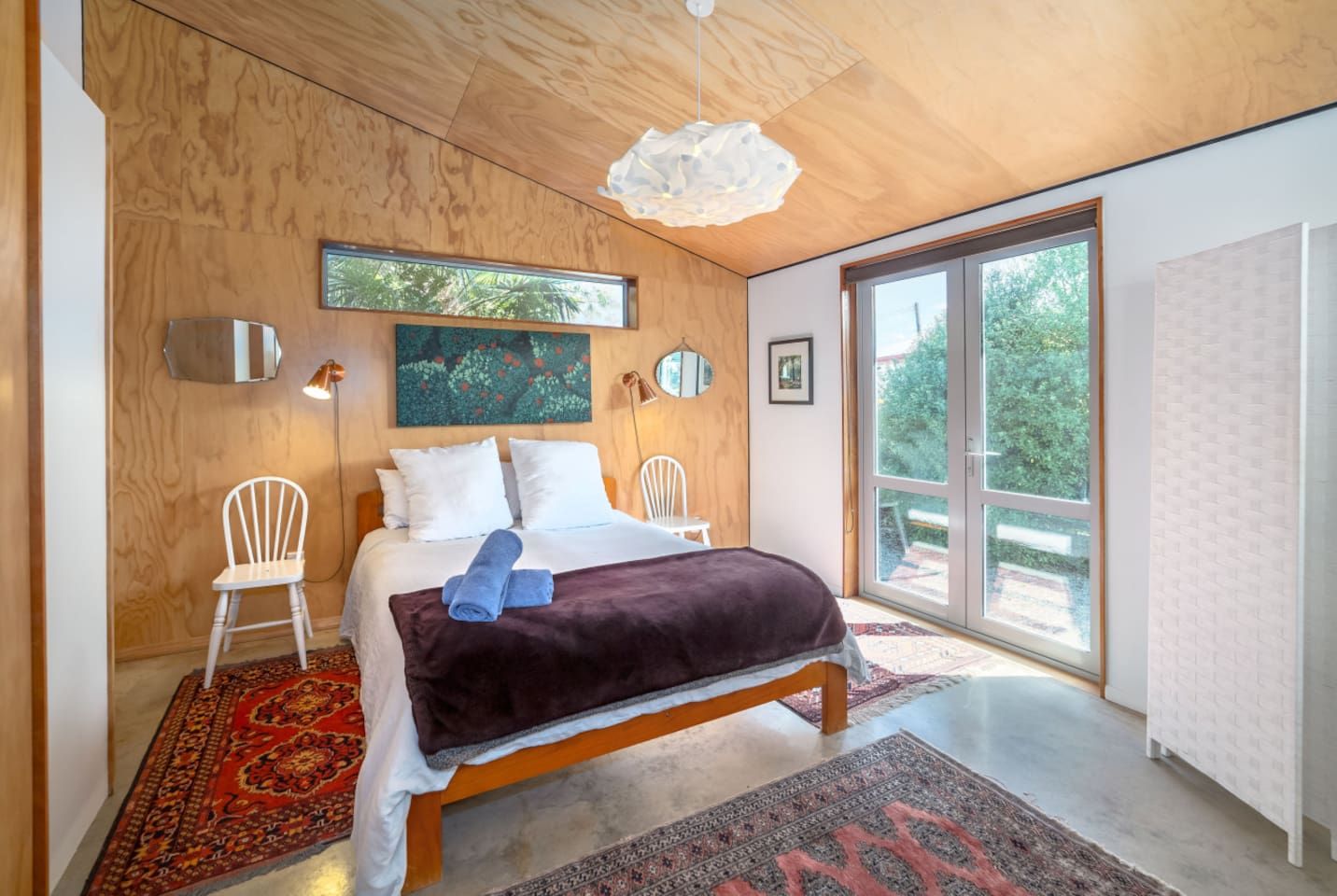 Un dormitorio moderno con una cama grande, dos sillones, techo de madera, un ventanal y vista a espacios verdes.