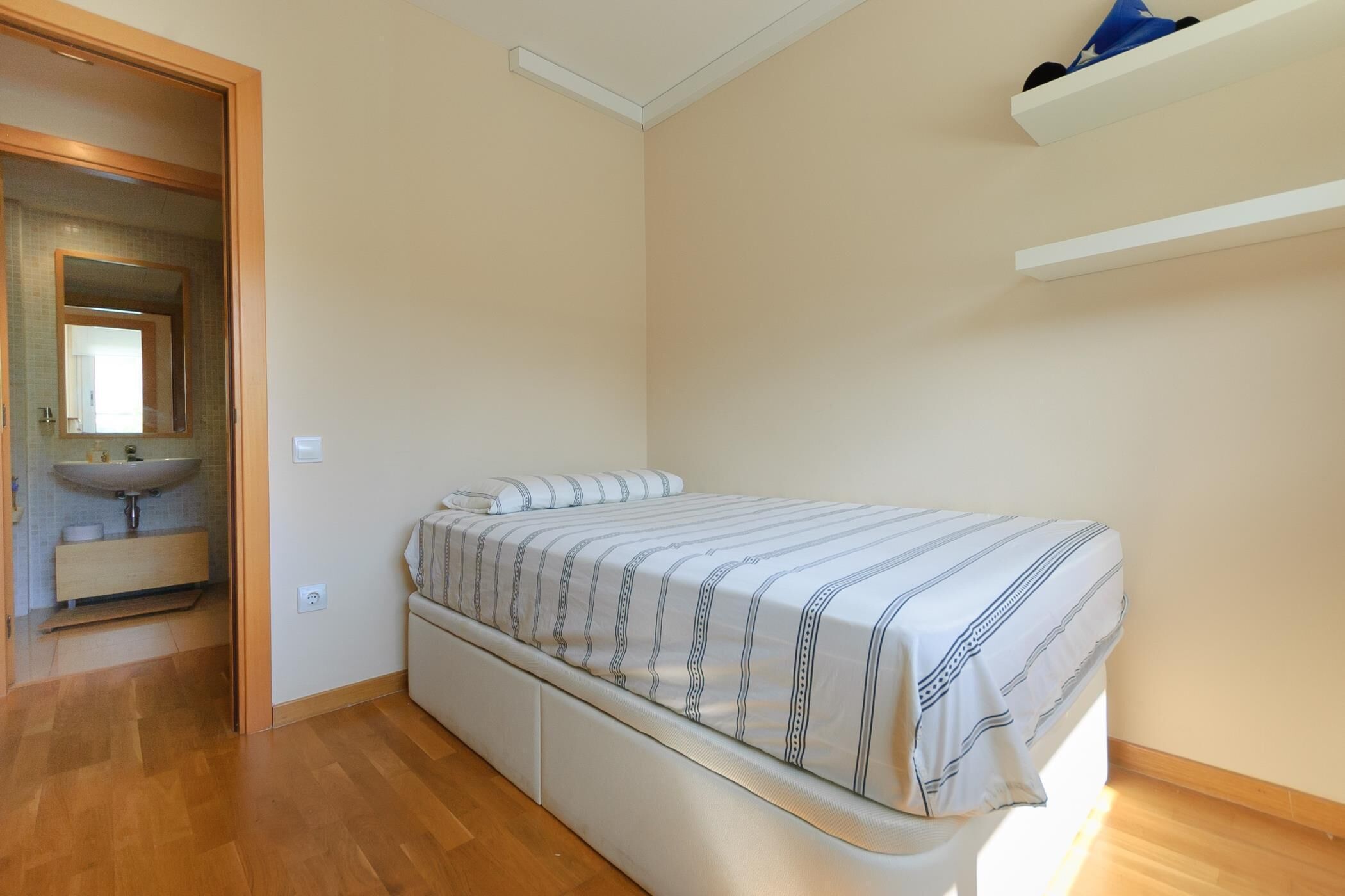 A bedroom with a bed, a built-in bench, a bedside table, a mirror, and a shelf above the bed.