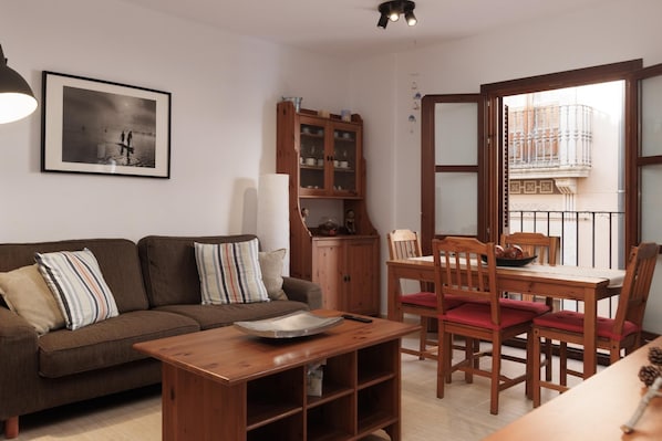 Una sala de estar con sofá, una mesa de comedor con sillas, un armario y un balcón con barandilla.