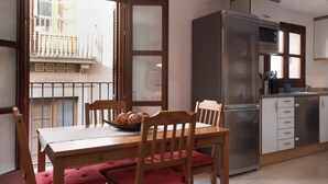 A dining area with a wooden table and chairs, a bowl of fruit, and a view of a balcony with a railing.