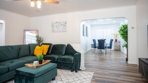 A modern living room with a green sectional sofa, a coffee table, and a ceiling fan. There is a dining area in the background with a table and chairs. - The Yuma White Modern Pool/Pet friendly Home (Yuma)