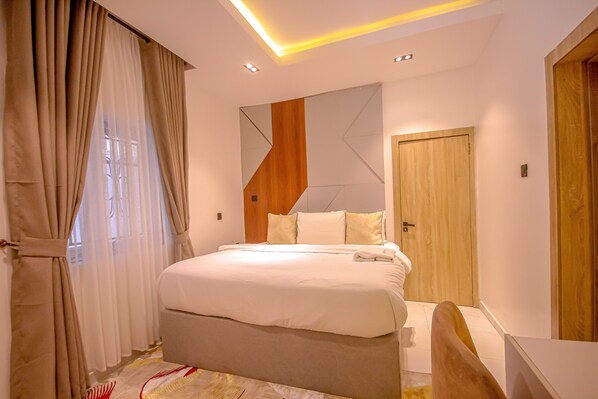A modern bedroom with a large bed, a wooden accent wall, and a wooden door. - Greyfield Apartments (Benin City)