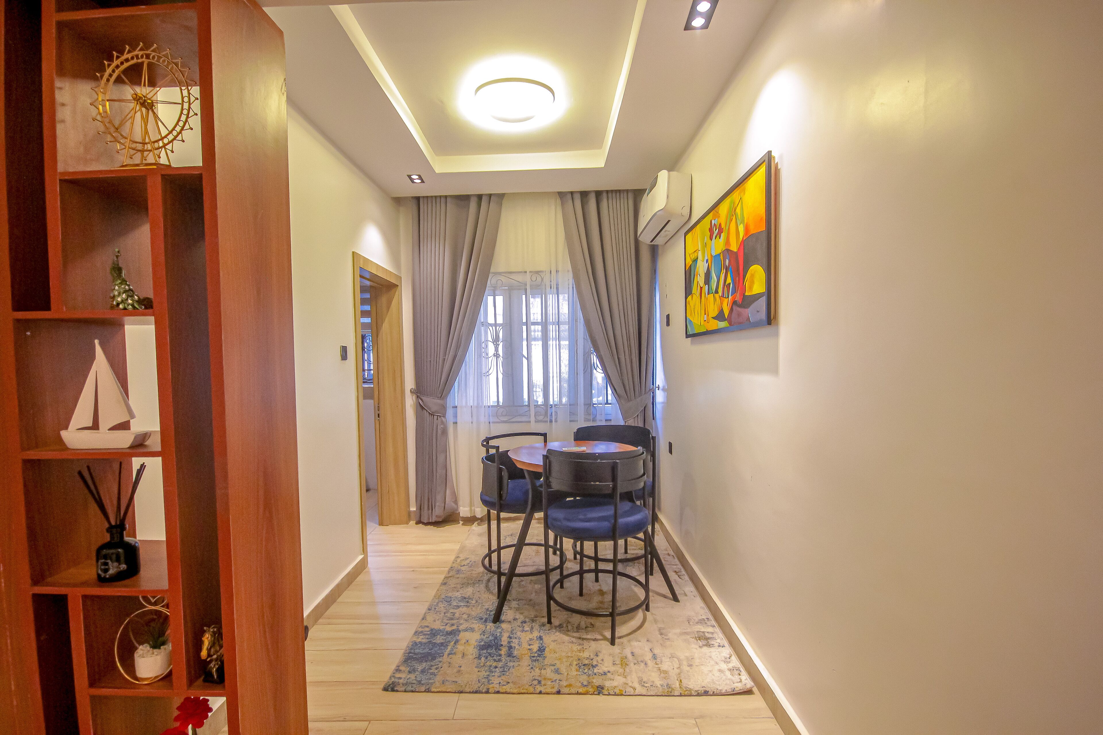 Um corredor com uma estante de madeira, uma área de refeições com mesa e cadeiras, uma luminária no teto e uma janela com cortinas.