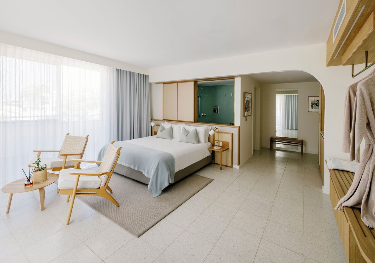 A modern hotel room with a bed, two chairs, a small table, a mirror, and a walk-in closet.