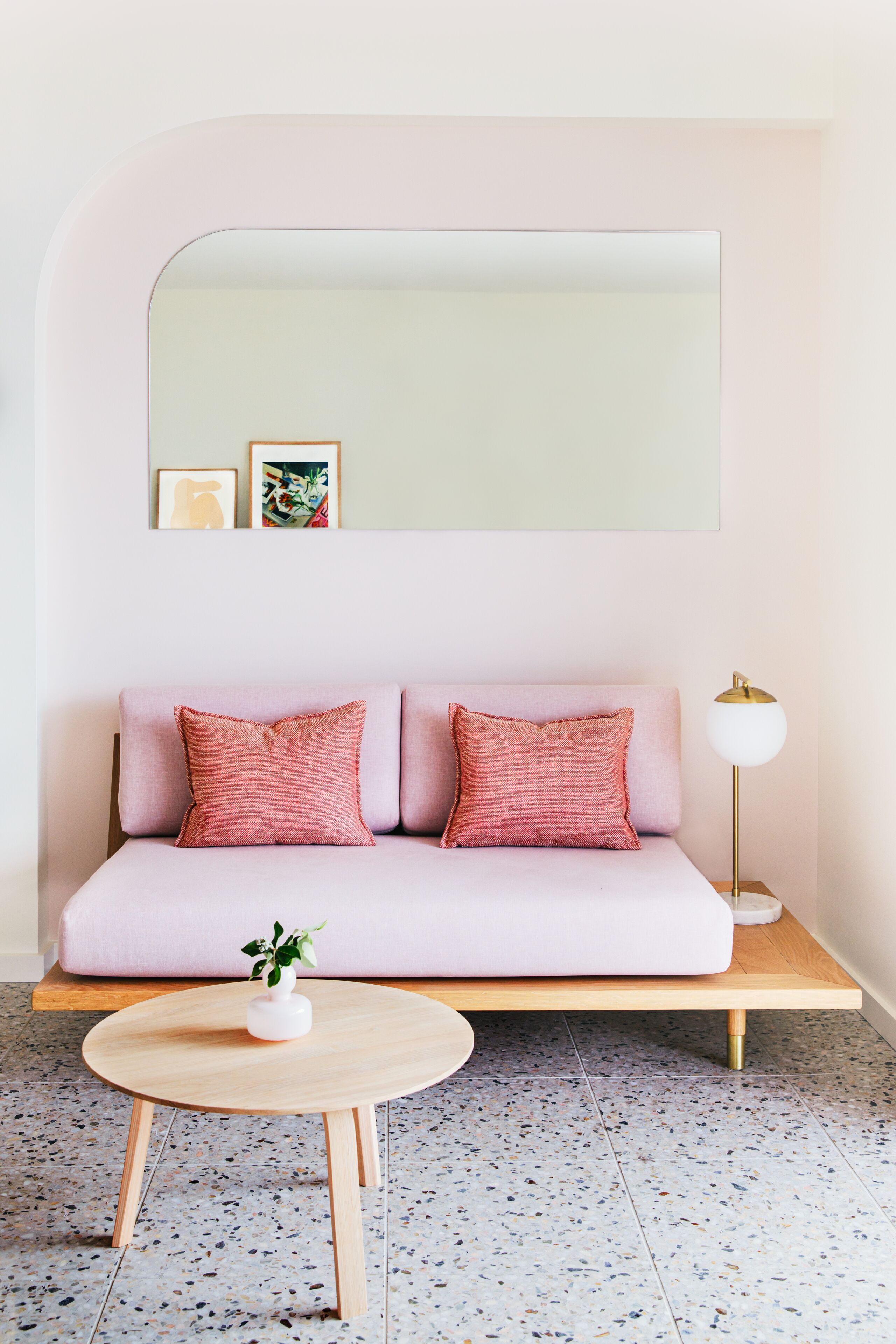 A room with a pink sofa, a round wooden table with a white vase, and a floor lamp.