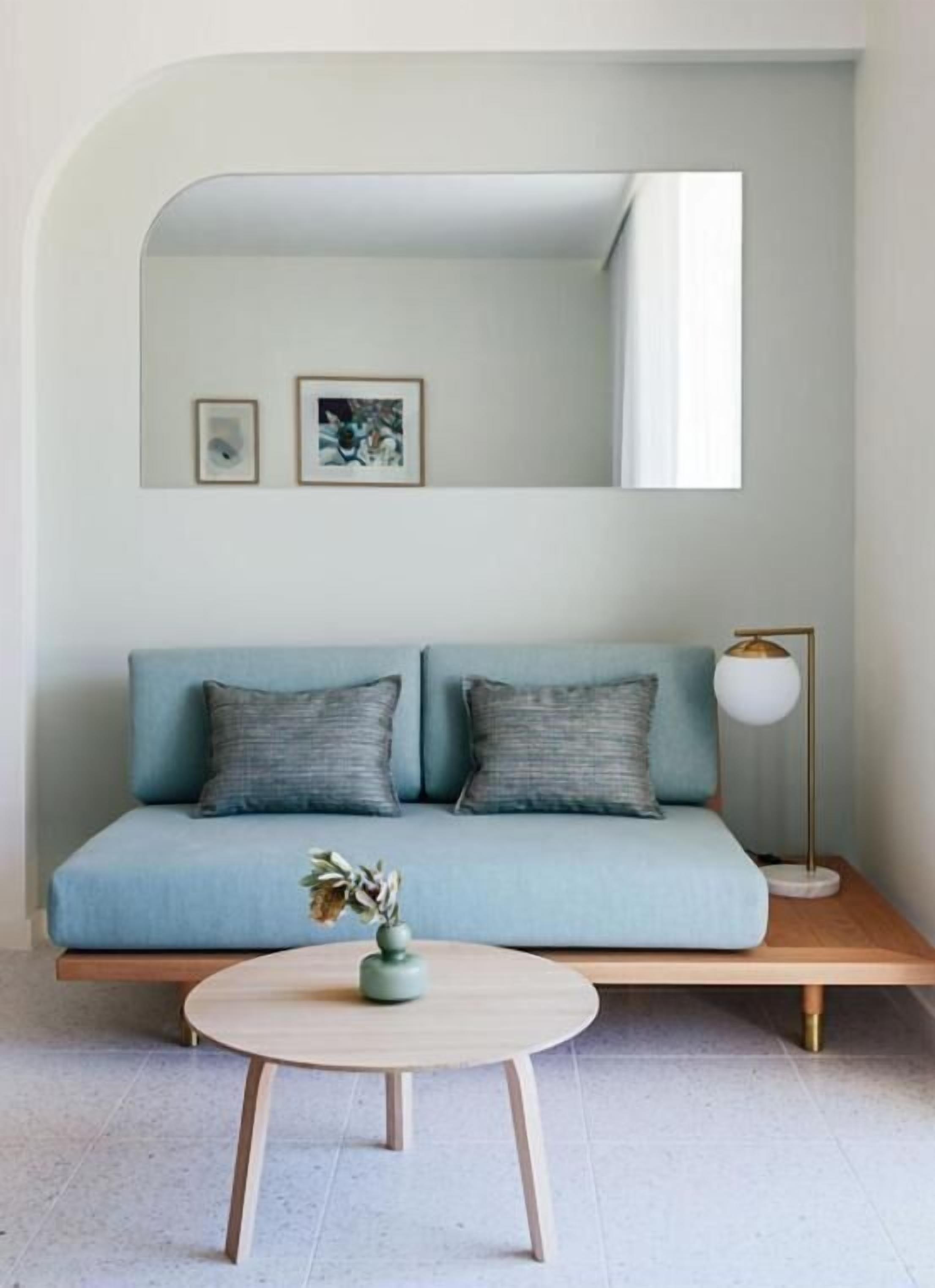 Un salon moderne avec un canapé bleu clair, deux coussins, une table basse ronde en bois sur laquelle est posé un vase, une lampe sur pied et un grand miroir au-dessus du canapé.
