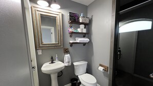 A bathroom with a pedestal sink, toilet, and a shower area. There are shelves above the toilet with toiletries and towels.